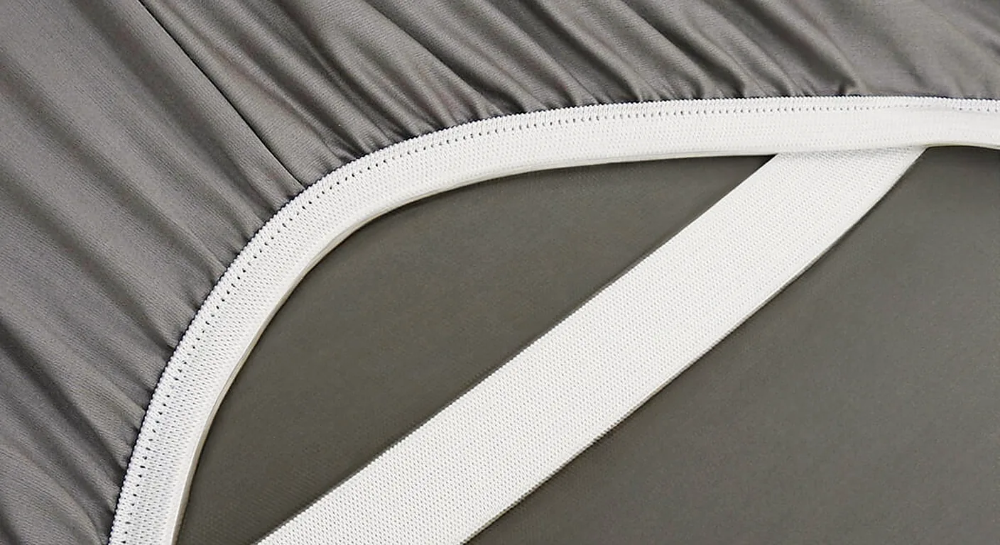 The underside of a grey fitted sheet corner with elastic all round and a white corner strap
